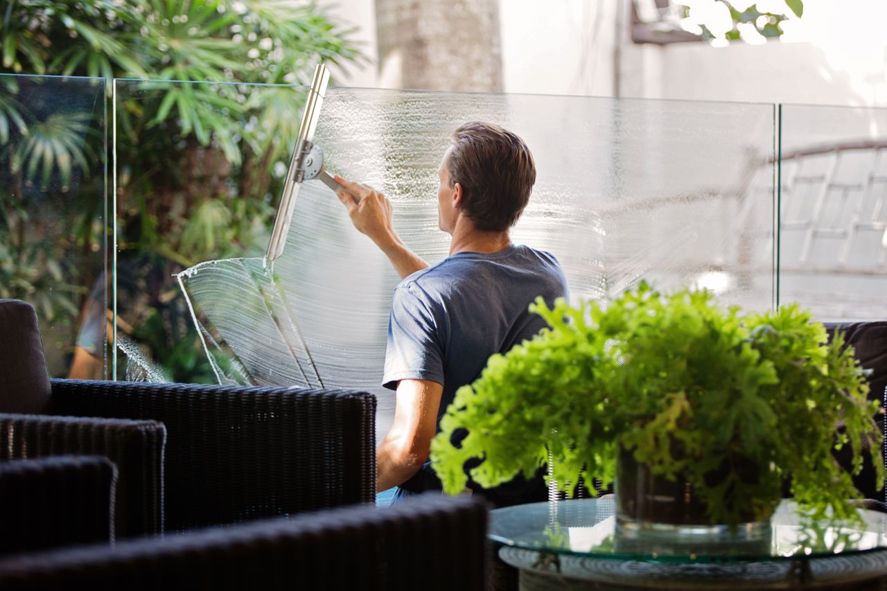 Un homme nettoie une barrière en verre à l’extérieur avec une raclette, entouré de végétation.
